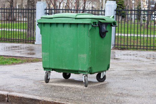 A green plastic wheeled rubbish bin with a textured surface, situated on a paved street near a curb. The bin has a closed lid with a slightly raised handle, and black wheels are visible at the base. It is positioned on a concrete surface with a wet appearance, suggesting recent rain. In the background, there is a black iron fence with vertical bars and decorative posts, enclosing a grassy area with trees and a pathway behind. The setting appears to be a residential or park area within Rickmansworth, with natural lighting indicating daytime. This image relates to waste management and rubbish removal services offered by Commercial Waste Rickmansworth in the local area.