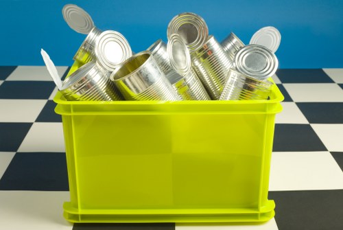 A bright yellow plastic bin filled with a variety of empty, unlabeled aluminium cans, some standing upright and others tilted or stacked haphazardly. The cans exhibit a metallic, reflective surface with ridged tops and are positioned in the foreground of the image. Behind the bin, there is a blue wall, and the bin is placed on a tiled surface with a black and white checkered pattern. The scene is well-lit, emphasizing the metallic sheen of the cans and the vibrant colour of the bin, suggesting an environment where waste collection or recycling activity related to aluminium cans takes place, consistent with rubbish removal services in Rickmansworth.