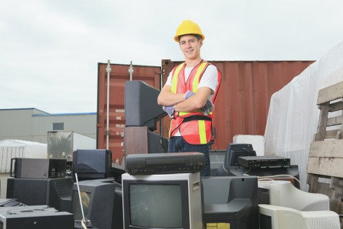 A young male worker at an outdoor waste disposal site in Rickmansworth, wearing a yellow safety helmet and high-visibility vest, is standing with arms crossed behind a collection of discarded electronic equipment, including old computer monitors, printers, and other obsolete electronic devices. The background features a large rust-colored shipping container, stacks of wooden pallets, and industrial warehouse structures under a cloudy sky. The electronic waste sits on the ground and on wooden pallets, with the worker appearing professional and prepared for rubbish removal tasks, representing a typical scene involved in commercial waste and electronic waste clearance services in the local area. The environment suggests an organized, outdoor storage or recycling yard dedicated to waste collection and disposal, aligned with the services offered by Commercial Waste Rickmansworth.